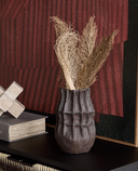A dark textured ceramic vase filled with dried grasses, displayed on a black shelf in front of a red and black geometric wall art piece.